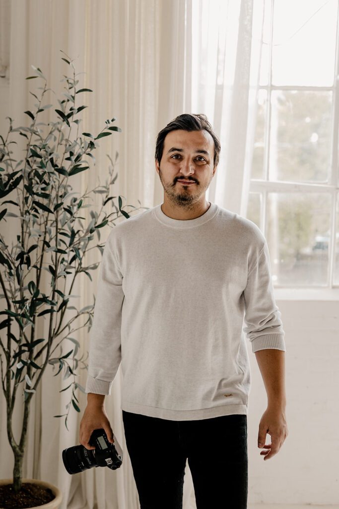 "A male photographer stands indoors, holding a camera in one hand, wearing a light sweater and dark pants, with a plant and large window in the background.