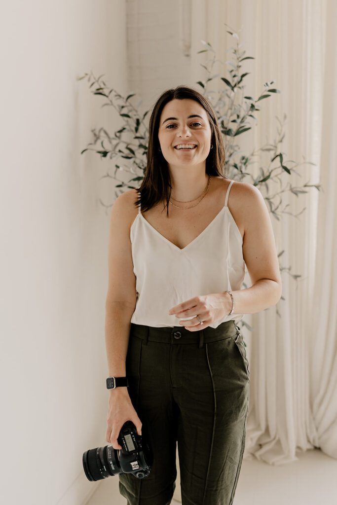 A female photographer stands indoors, smiling and holding a camera in one hand, wearing a white sleeveless top and dark pants, with a plant and white curtains in the background.Hevesi Journal  