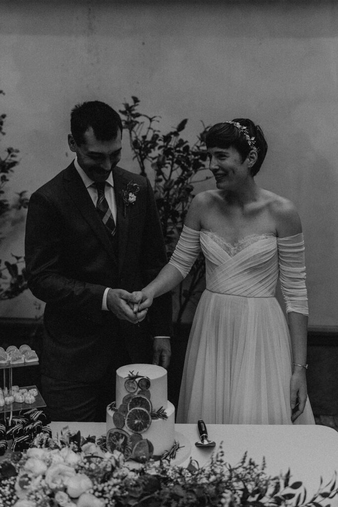 A bride and groom smiling as they cut their wedding cake together, with the cake beautifully decorated with fresh flowers and fruit.