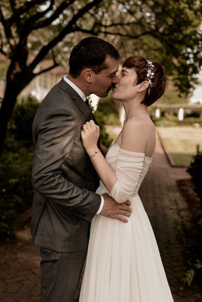 A bride and groom share a romantic kiss outdoors, with the bride holding the groom close and greenery in the background.