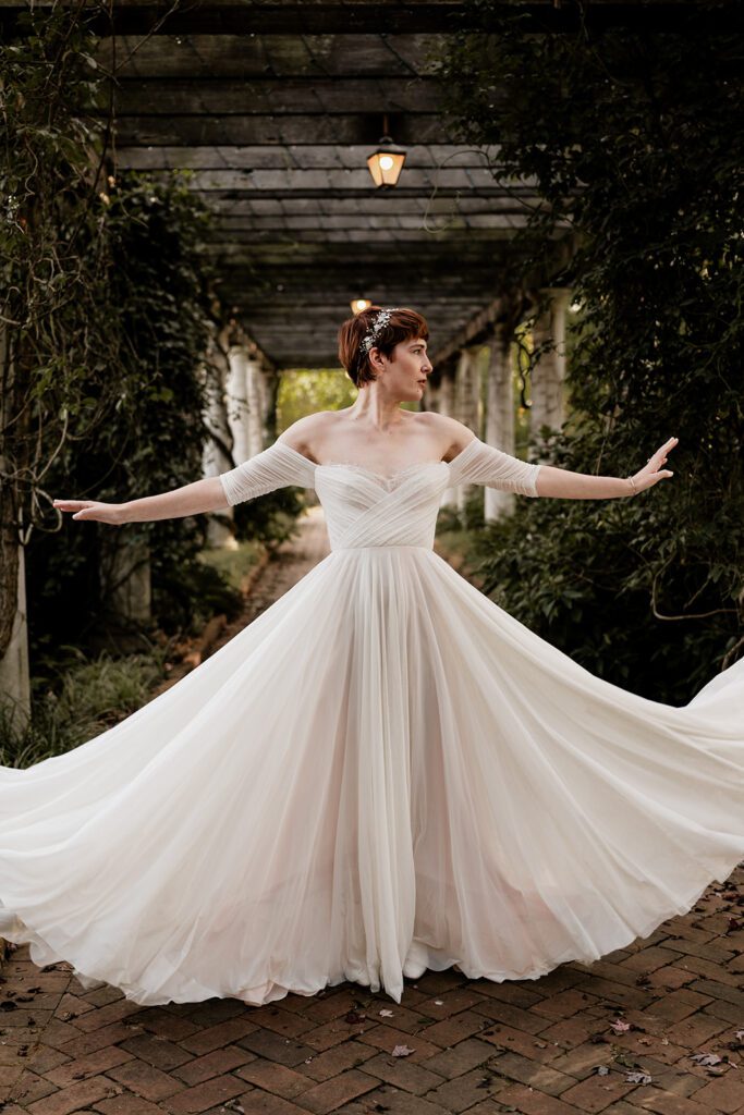 A bride twirls in her off-the-shoulder wedding dress, creating a beautiful flowing effect with her gown, standing under a vine-covered pergola.