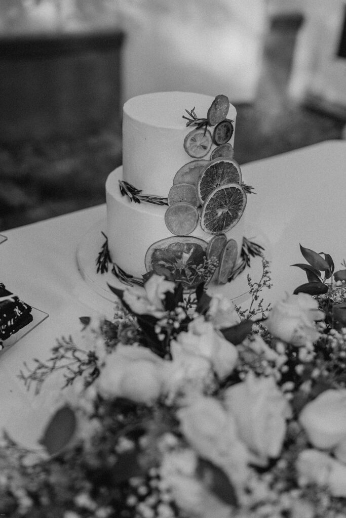 A black and white photo of a two-tiered wedding cake decorated with dried citrus slices and surrounded by floral arrangements.