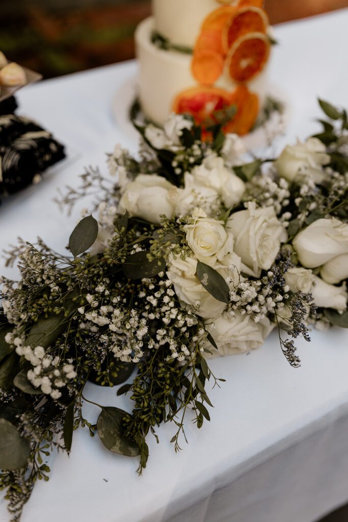 A close-up of a floral arrangement featuring white roses and greenery, placed next to a wedding cake decorated with dried citrus slices.