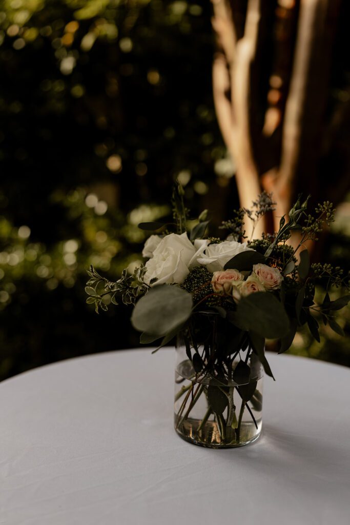 A floral arrangement in a glass vase placed on a white tablecloth-covered table, with greenery in the background.