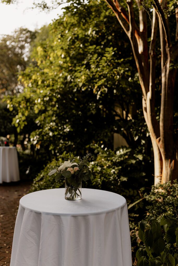 A round table with a white tablecloth and a small floral arrangement, set in an outdoor garden area.