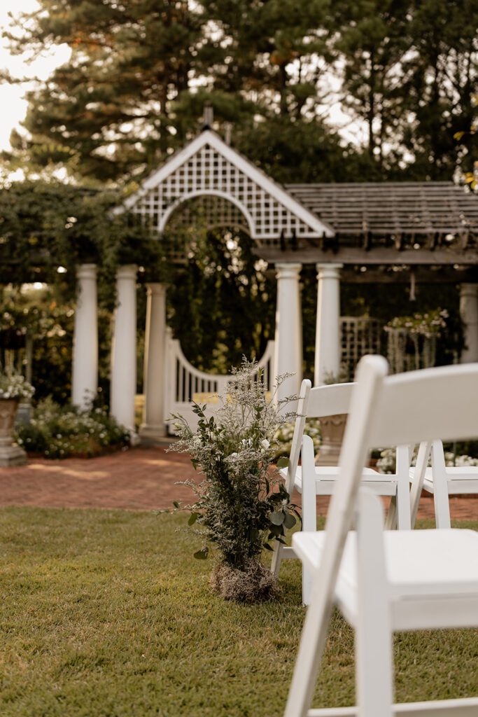 "An outdoor wedding ceremony setup with white chairs facing a white pergola covered in greenery.