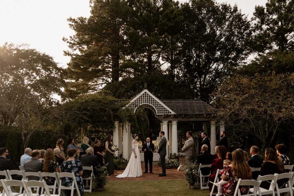 A wedding ceremony taking place on the Great Lawn, with the bride and groom standing under a white pergola surrounded by greenery, and guests seated in the foreground.