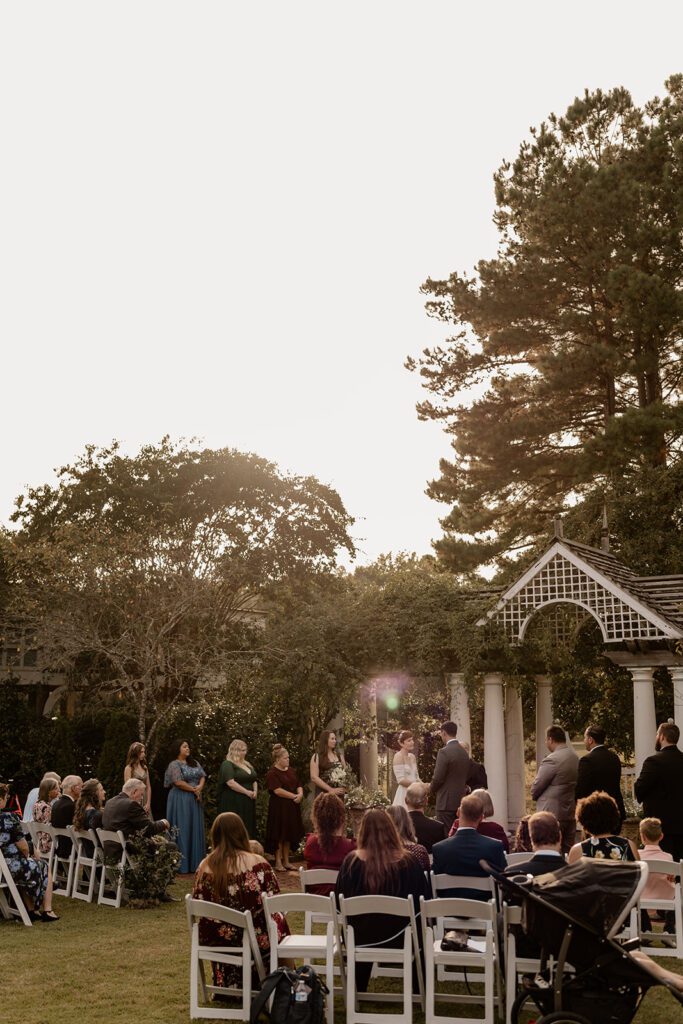 A wider view of a wedding ceremony on the Great Lawn, showing guests seated and standing, watching the bride and groom under a white pergola.