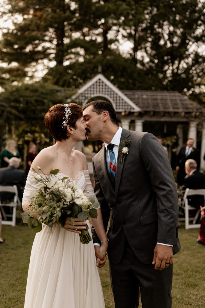 A bride and groom share a kiss after their wedding ceremony on the Great Lawn, with the bride holding a bouquet and the pergola visible in the background.