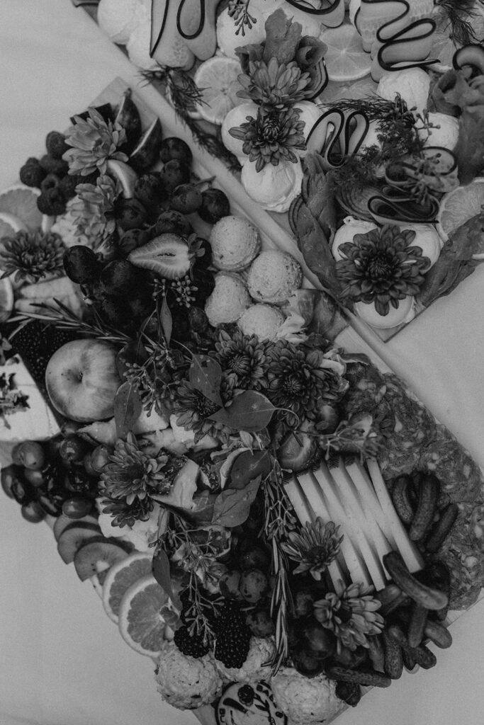 A black and white image of a beautifully arranged charcuterie board filled with a variety of fresh produce, meats, and cheeses, adorned with flowers.