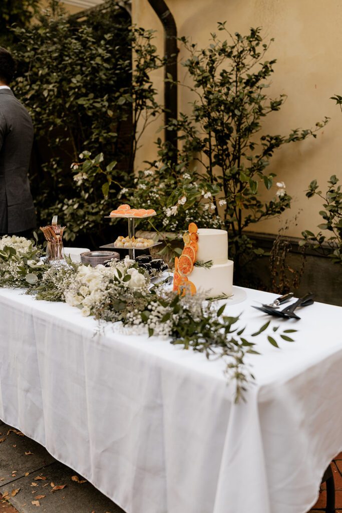 An elegantly decorated dessert table at an outdoor wedding, featuring a variety of cakes and desserts adorned with fresh flowers and greenery.