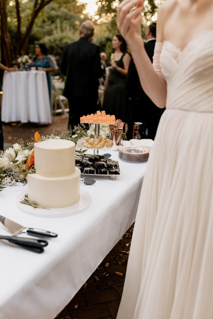 A close-up of a bride standing next to a dessert table, showcasing a beautifully decorated wedding cake and an assortment of desserts, with guests mingling in the background.