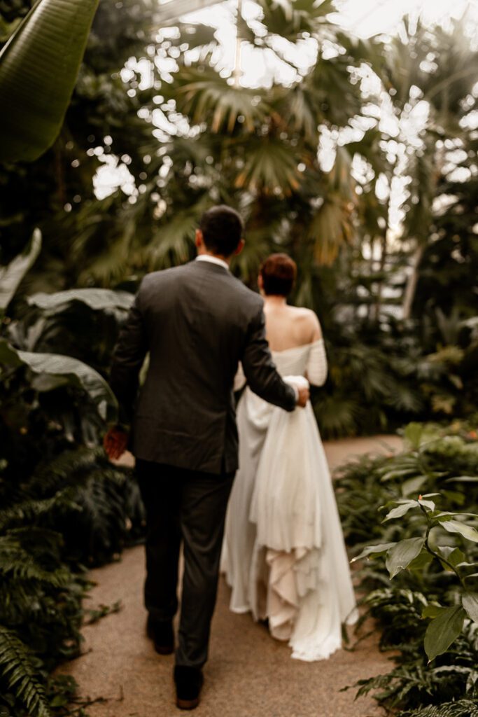 A bride and groom walk arm in arm through a lush, green pathway in the Orchid Garden, with the bride's dress trailing behind her.