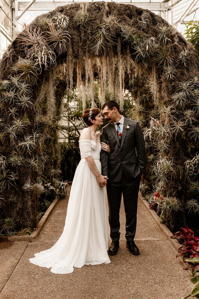 A bride and groom stand smiling under a lush, archway adorned with greenery and air plants in the Orchid Garden.