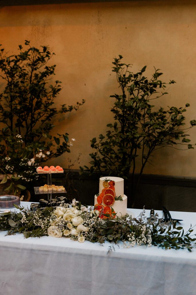 A beautifully decorated dessert table set up outdoors, featuring a wedding cake adorned with fresh flowers and fruit, surrounded by greenery.