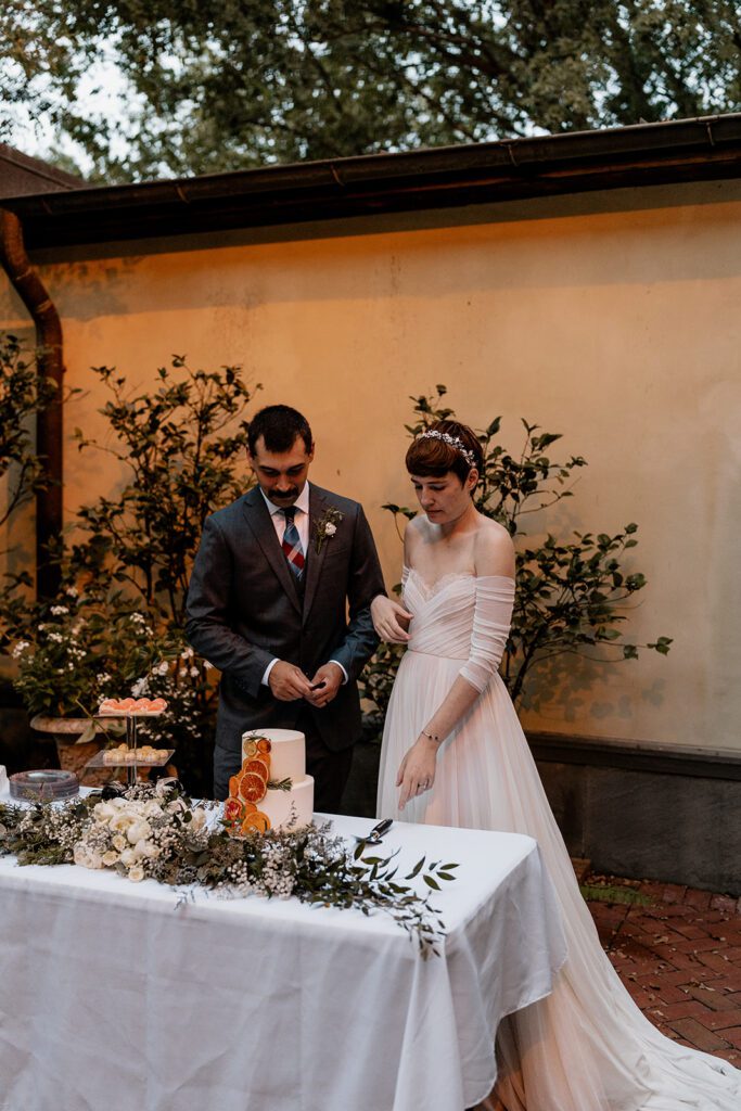 A bride and groom standing together at their wedding dessert table, preparing to cut the cake, with the cake and table elegantly decorated with fresh flowers and greenery.