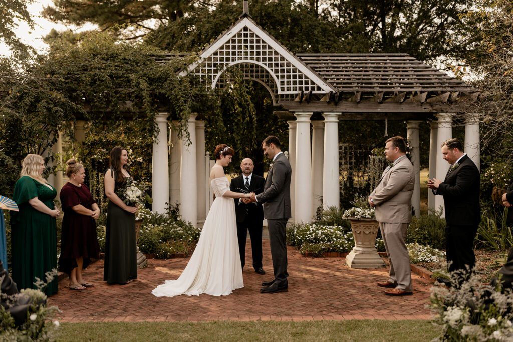 A bride and groom stand facing each other, holding hands during their outdoor wedding ceremony, surrounded by their wedding party under a white pergola draped with greenery.