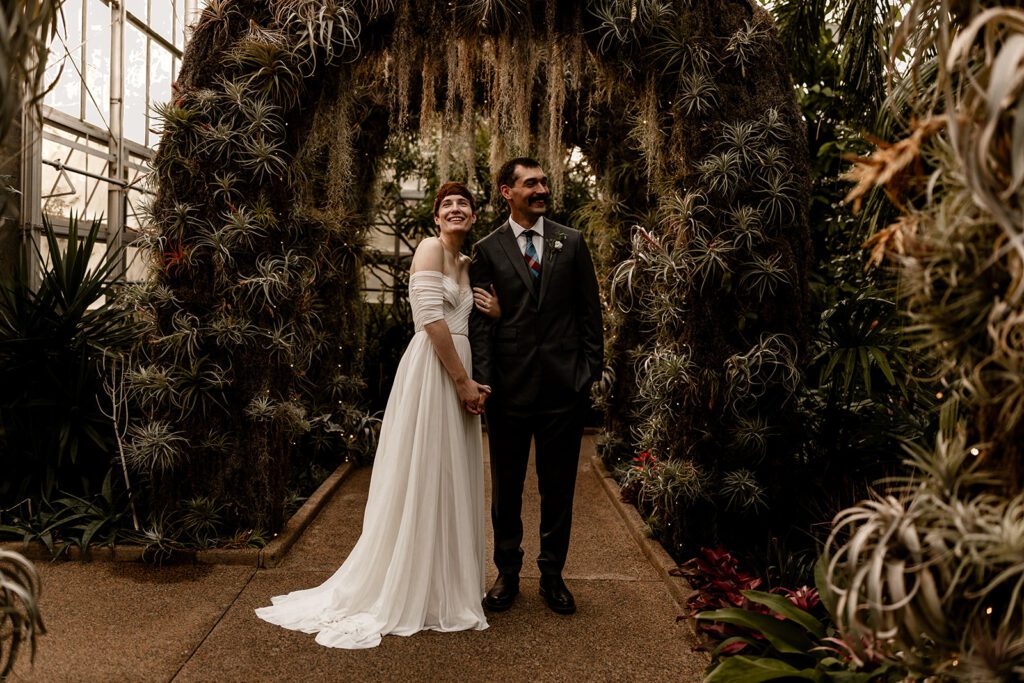 A bride and groom stand smiling under a lush, archway adorned with greenery and air plants at the Daniel Stowe Botanical Garden.