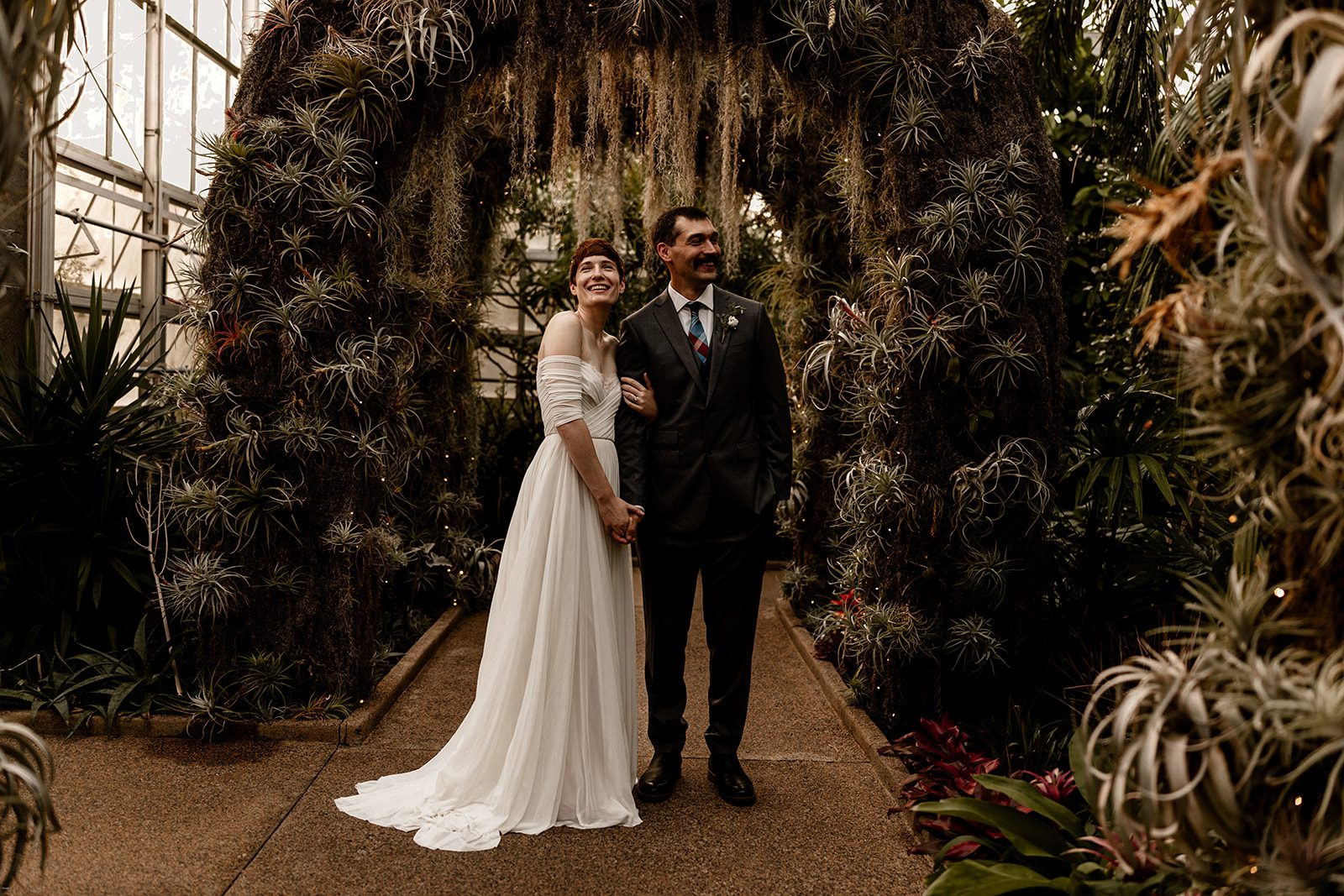 A smiling couple stands hand-in-hand under a lush, green archway covered with air plants and moss in a conservatory. The bride is wearing an off-the-shoulder white wedding gown, while the groom is in a dark suit with a plaid tie. They look off into the distance, surrounded by vibrant foliage.