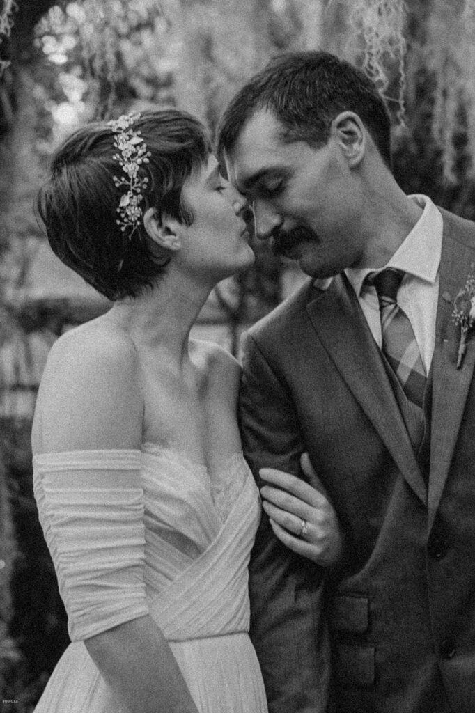 A bride and groom share a tender moment, smiling and leaning into each other, with the bride wearing an off-the-shoulder dress and floral headpiece and the groom in a suit and tie.