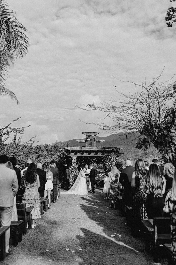 A black and white photograph of a wedding ceremony at Hacienda Siesta Alegre, with guests seated and standing, watching the bride and groom exchange vows under a floral-decorated pergola.