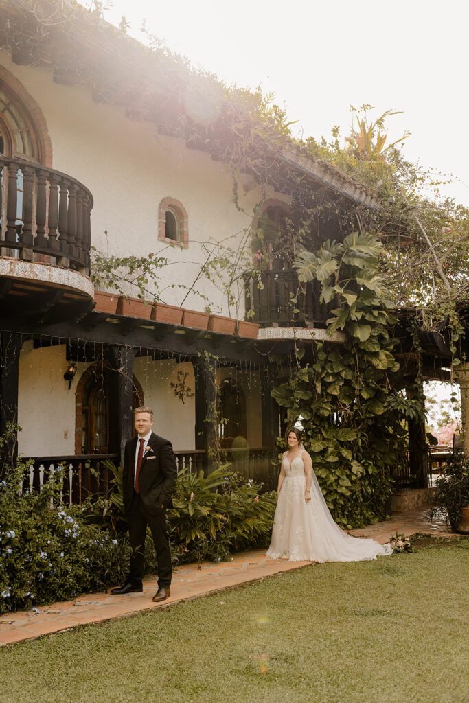 Bride and groom, Michael and Anna, standing in the garden area, surrounded by lush greenery and rustic architecture.