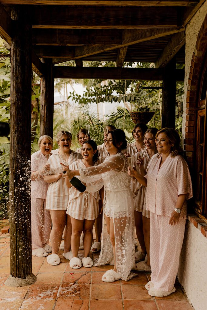 Bride, Anna, celebrating with her bridesmaids by popping a champagne bottle on a covered terrace, all wearing matching pajamas and slippers.