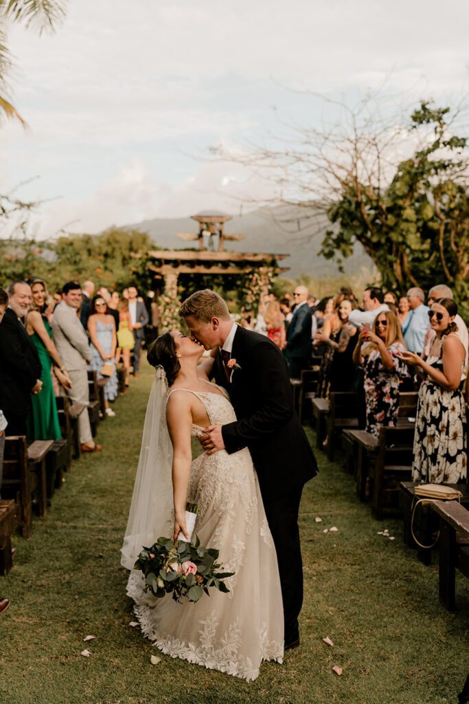 "A color photograph of a wedding ceremony at Hacienda Siesta Alegre, showing the bride and groom kissing as they walk down the aisle with guests applauding and smiling in the background.