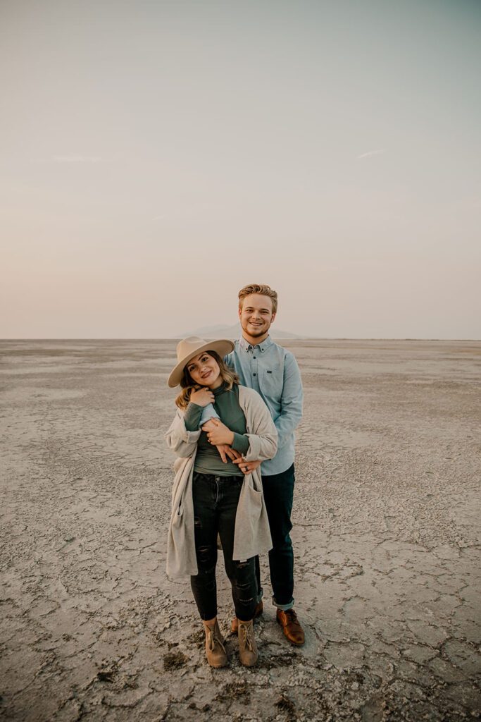 Couple embracing during their engagement photoshoot in a desert landscape, both wearing casual, coordinated outfits with a neutral color palette.