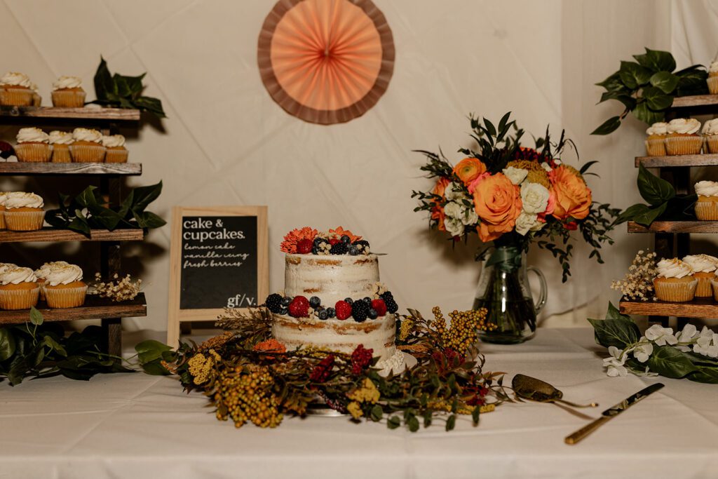 Rustic wedding dessert table with a semi-naked two-tier cake decorated with fresh berries and flowers, surrounded by greenery and floral accents. Display includes cupcakes, a floral arrangement with orange and white roses, and a small chalkboard menu listing dessert options.