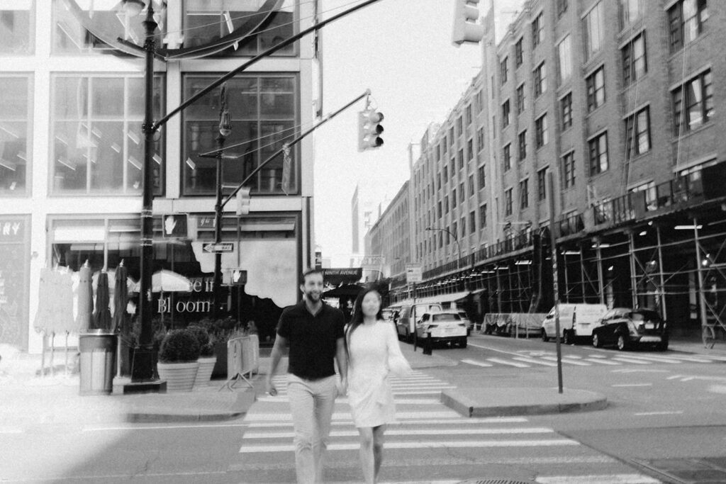 Black-and-white photo of engaged couple walking across the street in Chelsea NYC with brick buildings and storefronts in the background


