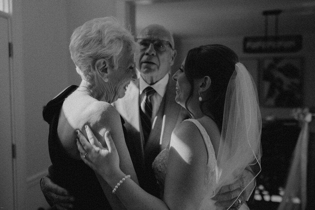 Bride embracing her grandmother while her grandfather looks on, captured in a candid black and white moment on the wedding morning.