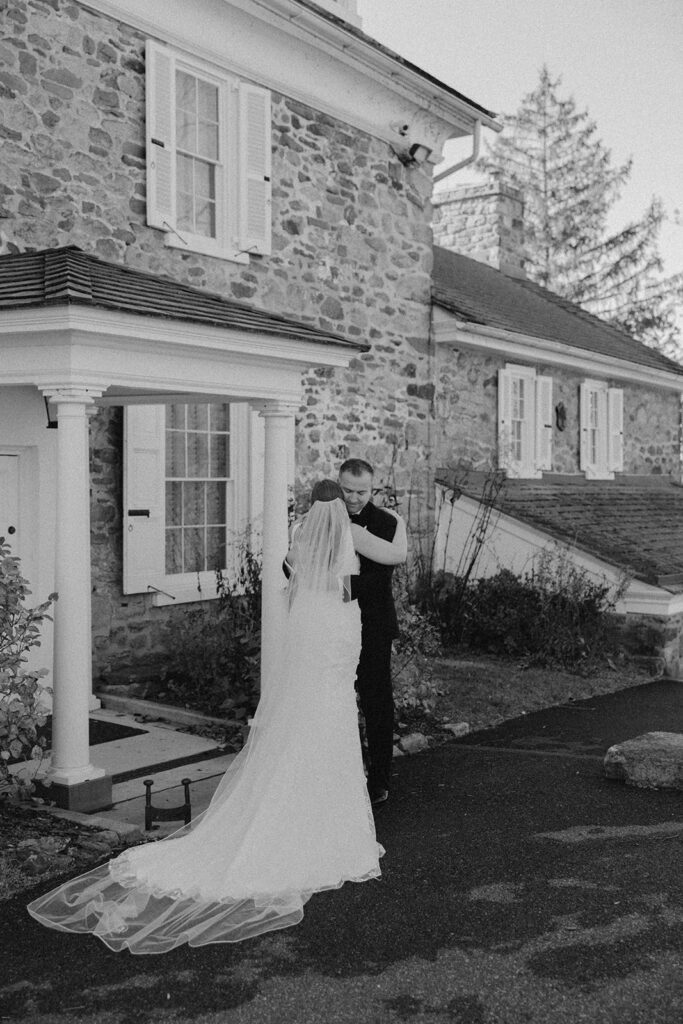 Bride and groom hugging beneath the porch of the stone house at the John James Audubon Center during a quiet black and white moment.