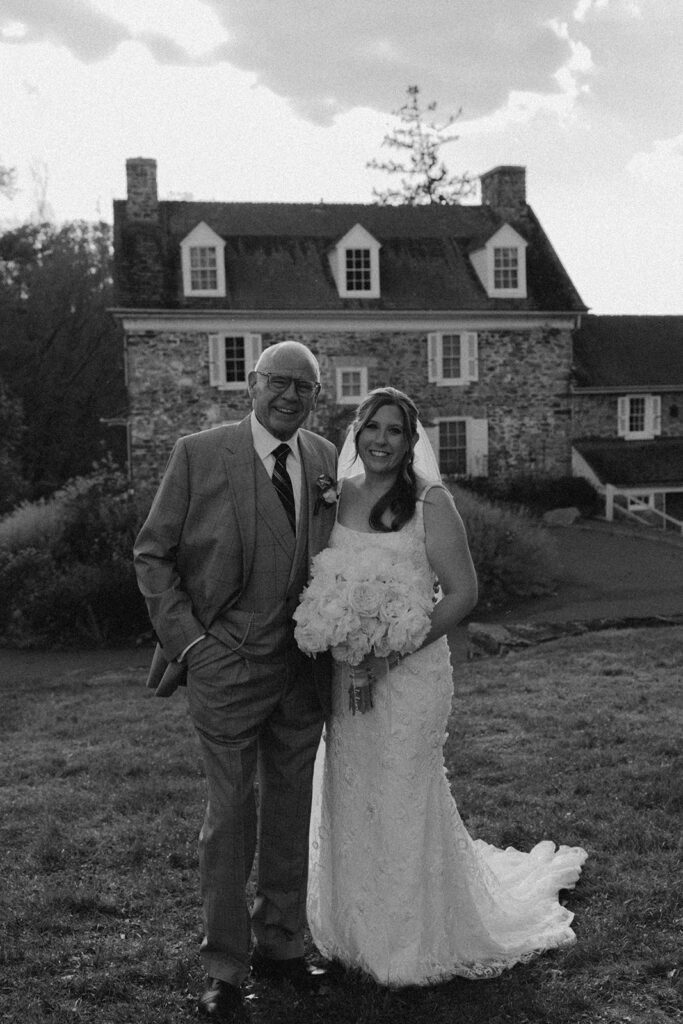 Bride standing with an older family member in front of the historic stone house at the John James Audubon Center, captured in a candid black and white portrait.
