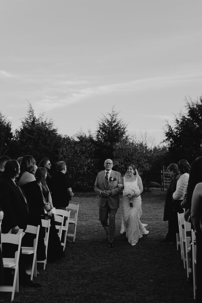 Bride walking down the outdoor aisle with her father as guests stand and watch during the ceremony.