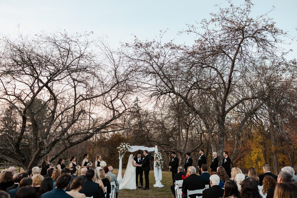 Bride and groom standing beneath the ceremony arbor in the orchard at the John James Audubon Center as guests look on.